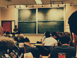 A teacher gestures at a chalkboard filled with mathematical equations