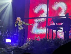 renee rap holidng a mic performing on stage with a red screen displaying the number 23 in white. She is wearing a green long sleeve and black pants. To the right of her is a man playing the keyboard